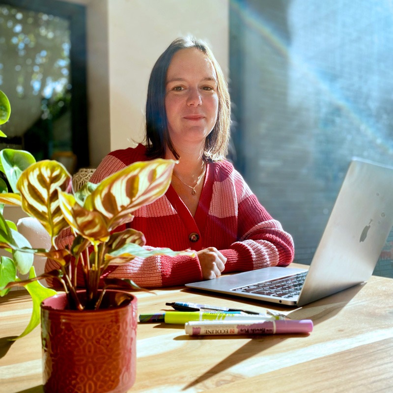 Portrait de Sophie Bortier, assise à son bureau avec son ordinateur portable devant elle, jetant un regard confiant et chaleureux vers l'objectif. L'arrière-plan est sobre, mettant l'accent sur son espace de travail.